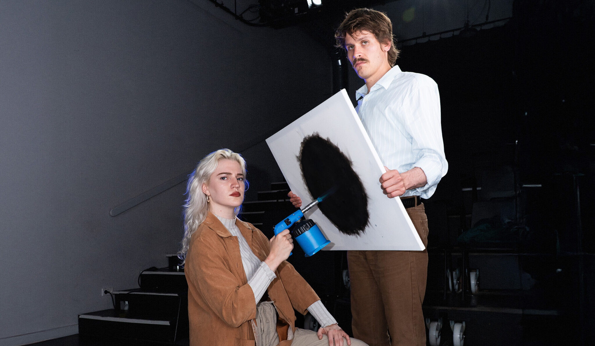 vrouw maakt met een brander een zwarte cirkel op een canvas, vastgehouden door een man, campagnebeeld voor voorstelling NUL