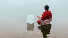 Campagnebeeld van de voorstelling Met de stroming mee. Je ziet een vrouw in het water staan tot haar heupen met een drijvende jerrycan naast zich.