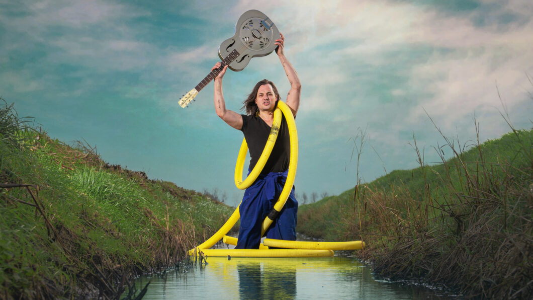 Dit Verdronken Land - man in een sloot houdt een gitaar boven zijn hoofd