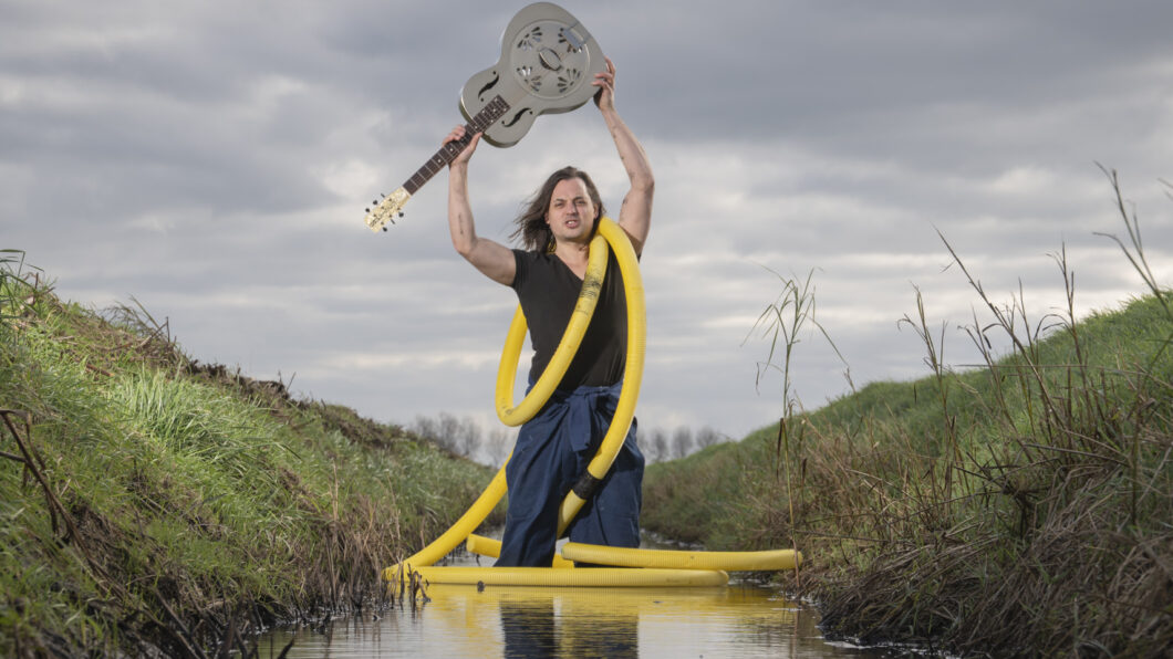 Dit Verdronken Land - man in een sloot houdt boven zijn hoofd een gitaar in de hand