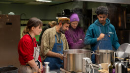LACUNA KITCHEN: Vier mensen staan in een professionele keuken samen te koken achter een fornuis met grote, stomende pannen. Ze dragen schorten en kijken geconcentreerd in de pannen terwijl ze roeren en proeven; op het aanrecht staan glazen, flessen en kookgerei.