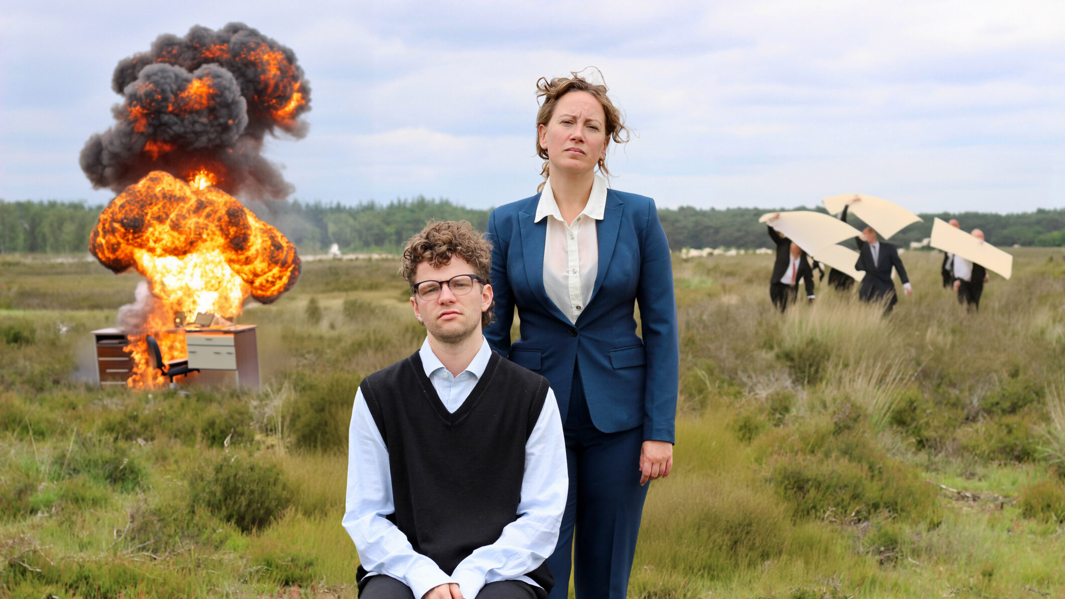 Twee serieus kijkende mensen in nette kleding staan en zitten in een open veld, terwijl achter hen een bureau in brand staat en een grote explosie plaatsvindt; op de achtergrond lopen anderen weg met panelen boven hun hoofd.