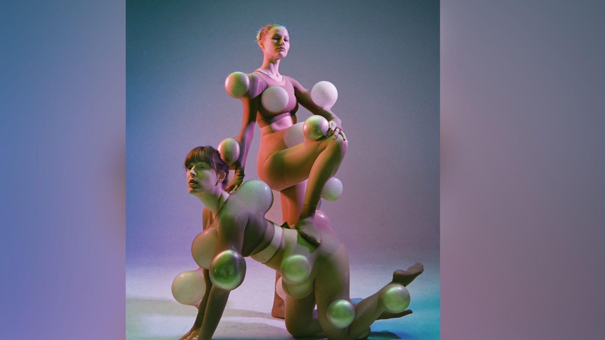 Twee performers poseren in een studio met zachte paarse en groene belichting. Ze dragen huidkleurige kostuums met ronde, bolvormige objecten die op verschillende plekken aan hun lichaam zijn bevestigd, waardoor hun silhouetten vervormd en overdreven worden. Eén performer knielt op handen en knieën terwijl de ander met één voet op haar rug staat. De afbeelding speelt met vormen, lichaamsbeelden en sculpturale poses. Campagnebeeld van Pretty Privilege