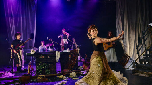 Voor jou, van mij: Een theatraal optreden op een donker verlichte scène met paarse spots. Op de voorgrond danst een vrouw in een lange, beige rok en een zwart top, met haar armen elegant uitgestrekt. Op de achtergrond speelt een band bestaande uit een gitarist, een drummer, een violiste en een gitariste. De scène is aangekleed met verspreide boeken, bloemen en gordijnen, wat een chaotische, creatieve sfeer creëert.